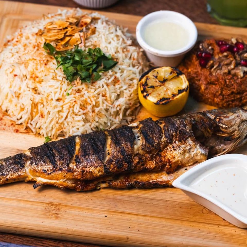 A Mediterranean grilled whole fish with rice, grilled lemon, and dips on a wooden board.