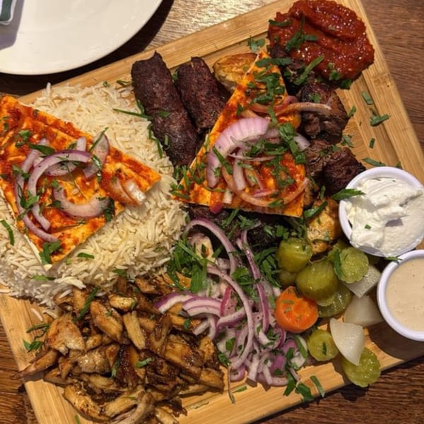A large Mediterranean platter with rice, various grilled meats, flatbread, pickled vegetables, and dips.
