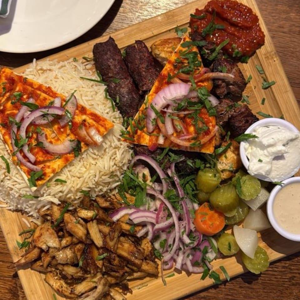 A large Mediterranean platter with rice, various grilled meats, flatbread, pickled vegetables, and dips.